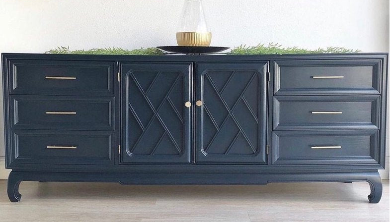 A dark navy blue wooden sideboard or credenza with curved legs, featuring two central cabinet doors with an X-pattern and four drawers on each side with slim gold pull handles. It is styled with greenery and a glass vase on top against a white wall and light wood floor.